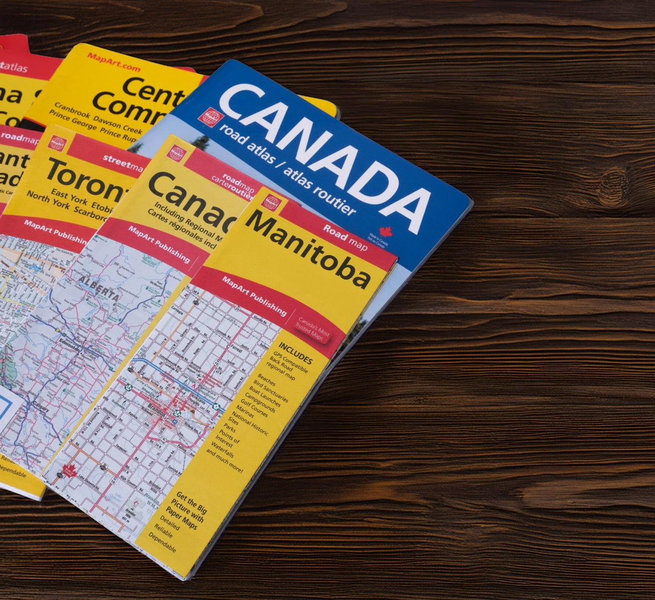 A Canada road atlas map book titled 'Manitoba' lies on a dark wood surface, partially overlapping other atlas volumes including 'Toronto' and 'Centra'. The cover features a blue top section with 'CANADA' in large white letters and a yellow lower section with red accents and map details.